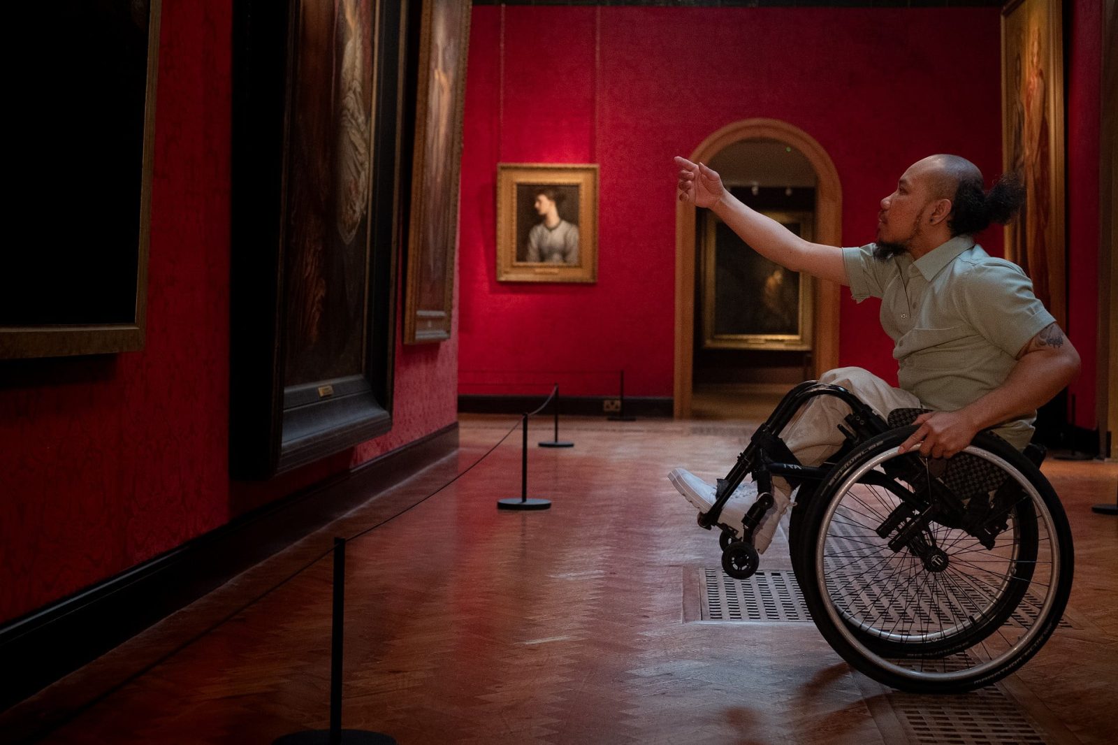 A solo dancer, performing seated in his wheelchair, his fore arms close together and reaching upwards. The gallery in which he performs is painted a deep red, and filled with pictures in golden frames.