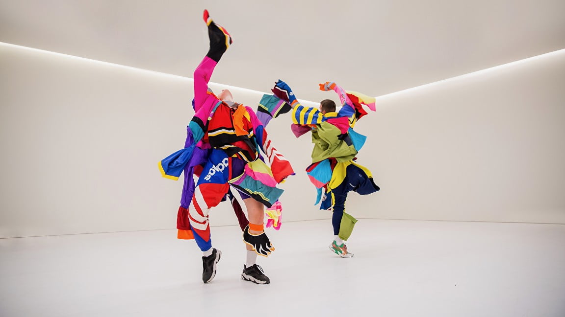 2 dancers wearing lots and lots of layers of multi-coloured clothes dance freely with their arms reaching up to the sky and their heads thrown back. They dance in a white cube gallery space with a single strip of fluorescent light all around the wall.