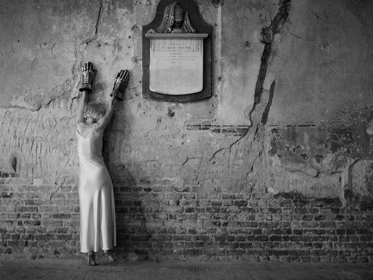 A lady in a long white dress made from a thin, shiny fabric leans against the crumbling stone wall of what looks like an old church, her arms extended above her head and her face concealed from the camera. She wears large metallic medieval gloves. The picture is black and white which emphasises the range of different textures, of the crumbling wall, its cracks, her heavy metallic gloves, the softness of her dress.
