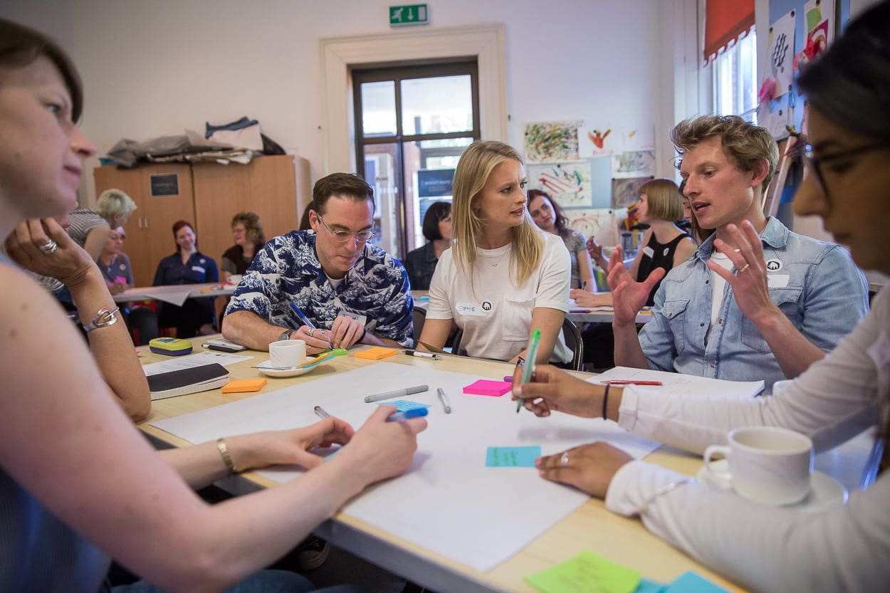 A group of people are seated around a table in a museum workshop room, with the table spread with large pieces of paper, pens and post-its. They listen intently to a man on the right side of the image as he describes something.