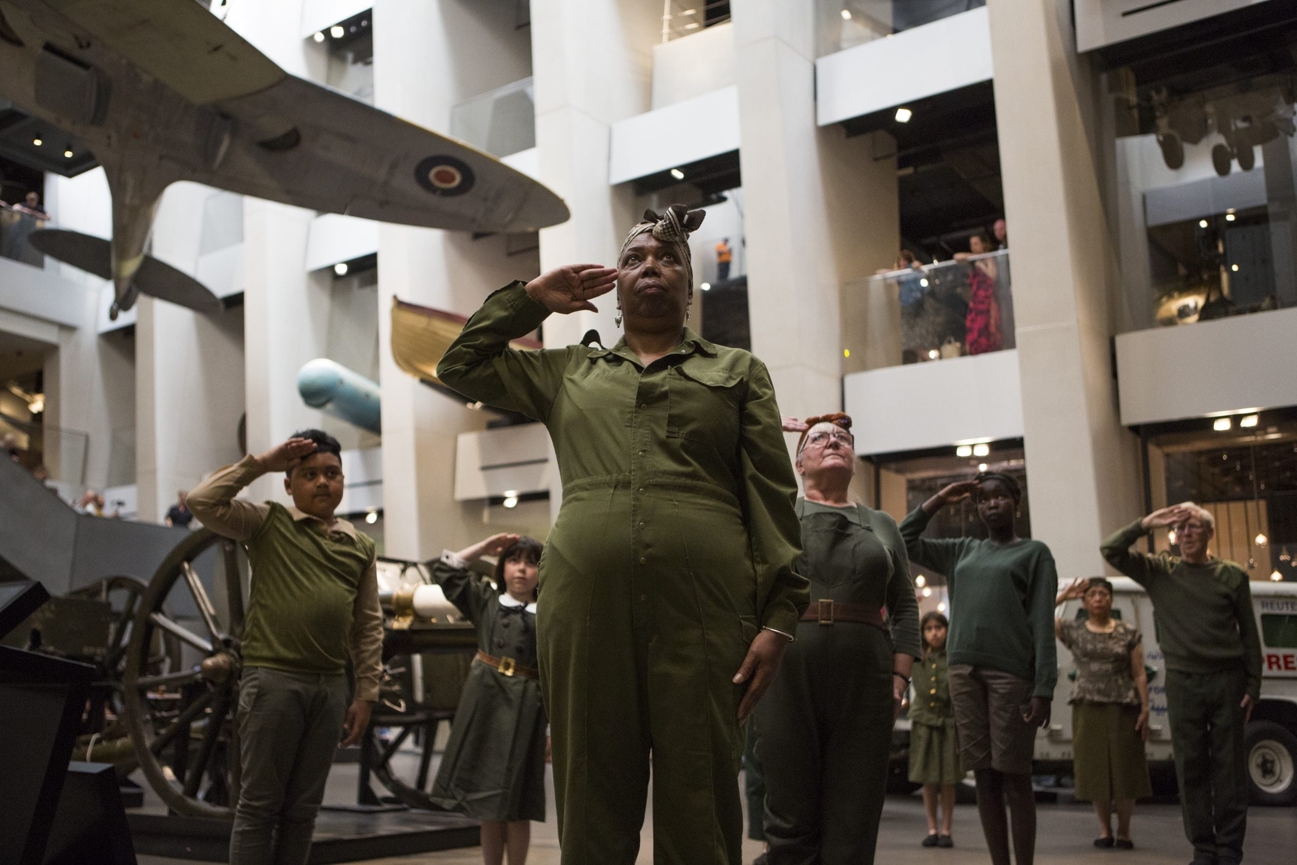 A group of dancers of all ages stand to attention, saluting with their right hands at their brows. They are part of a performance at an Imperial War Museum, and are dancing in front of planes in the exhibition.