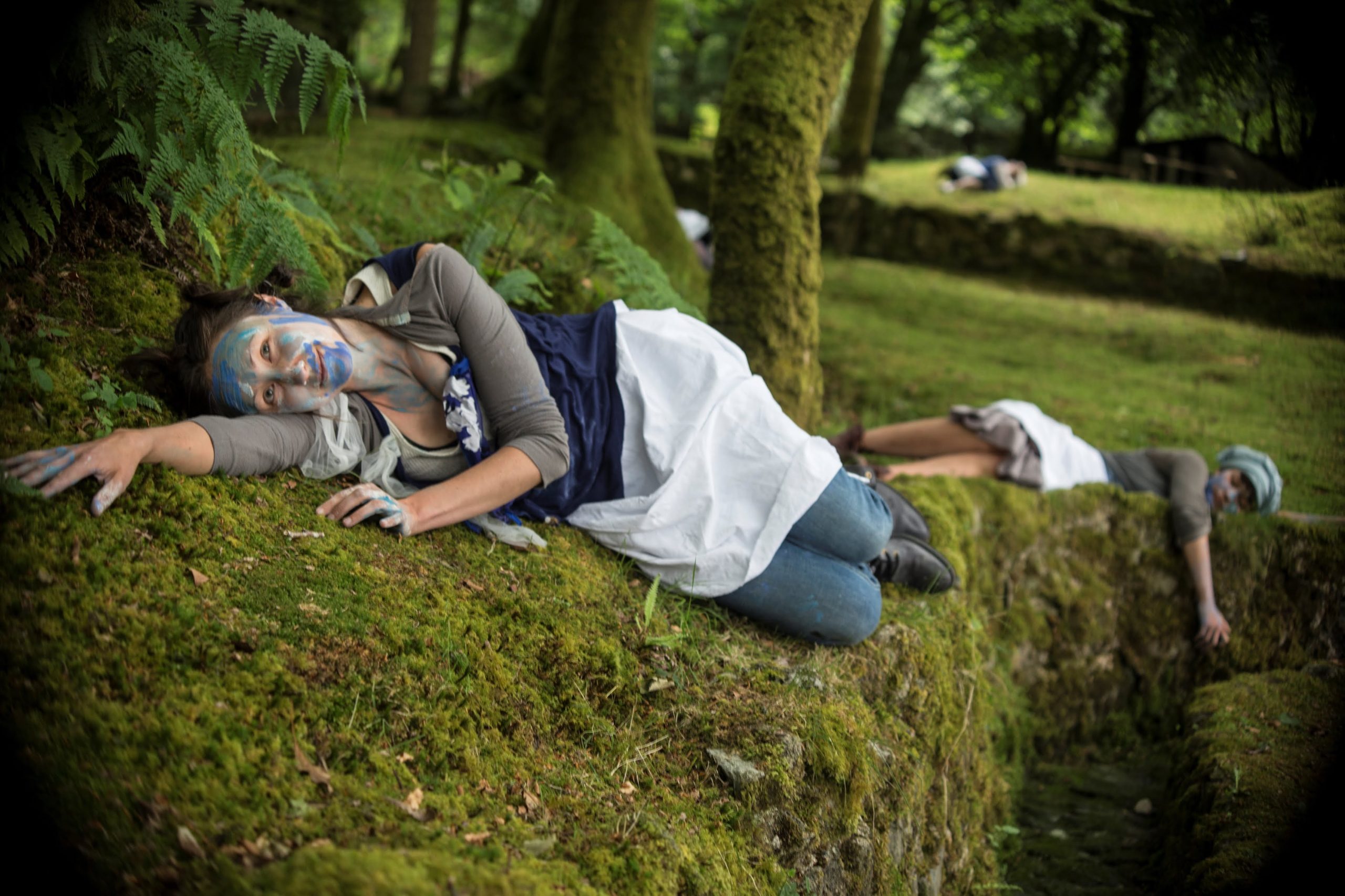 Young dancers, wearing costumes of blue and white, lie on their sides amongst the green woodland landscape of Wheal Martyn Clay Works in Cornwall
