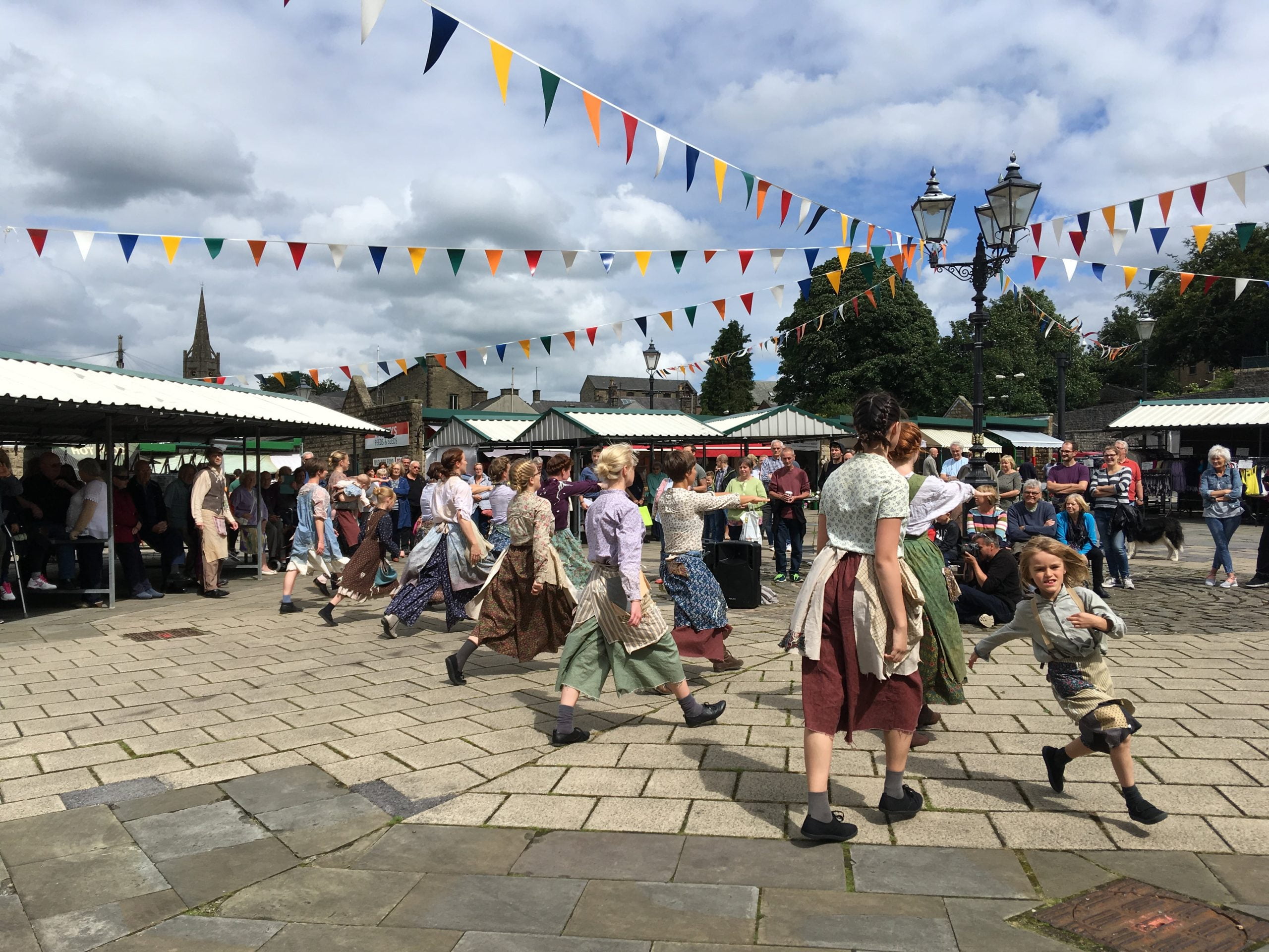 A group of dancers of all ages come together to dance in a town square in North West England. Colourful bunting hangs above their heads.