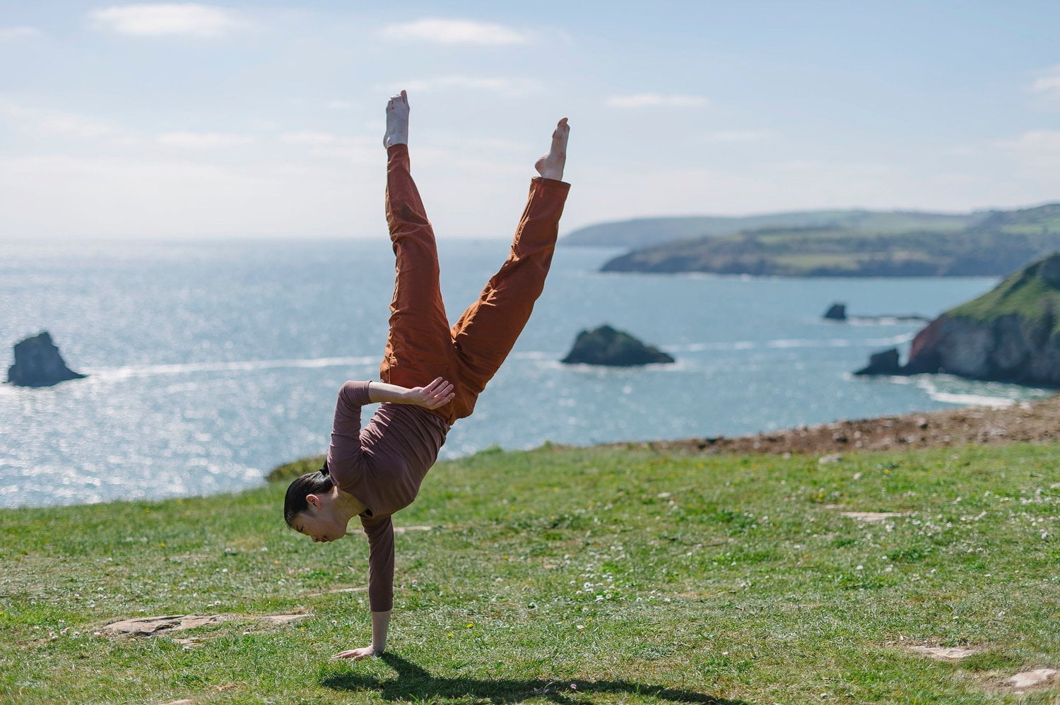 A solo dancer performs a one-handed handstand on the grass, a few metres from a cliff edge. Beyond her we can see the sea, and pieces of land jutting out into the blue water.