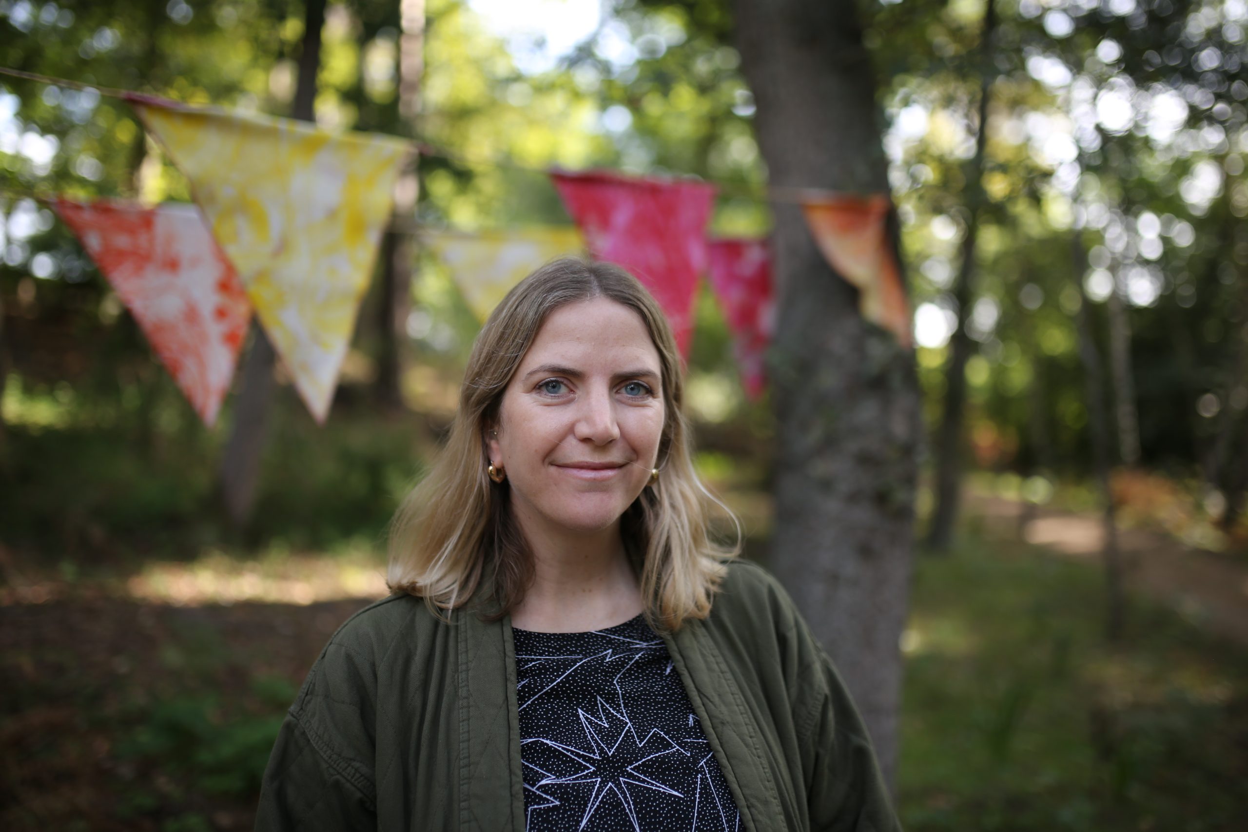 A picture of a white woman with brown hair to her shoulders standing in the grounds of Watts Gallery Artists' Village. She wears a blue top and dark green cardigan and there is red, yellow and orange bunting hung in the trees behind her.