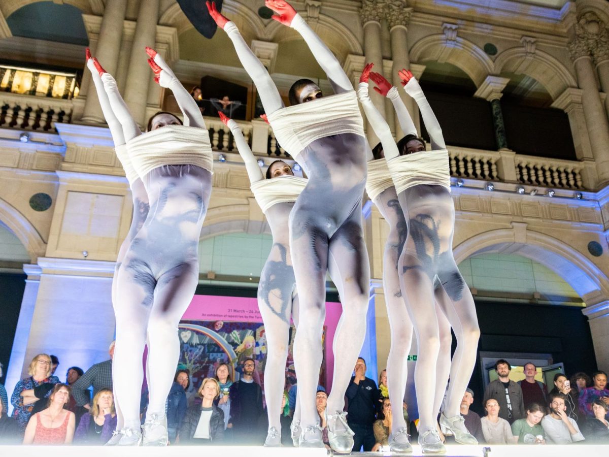 A group of 5 dancers perform on a catwalk set up in the grand opening hall of the Bristol Museum. They wear long transparent white unitards with black markings underneath and the tips of their hands are painted red. Their arms are stretched over their heads and some of the transparent white fabric is pulled up to cover their faces.