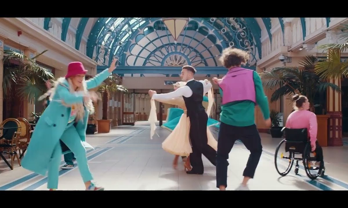 A group of different dancers, using different dance styles, all wearing bright blues, pinks, black and white dance under the blue arches of a historical building.