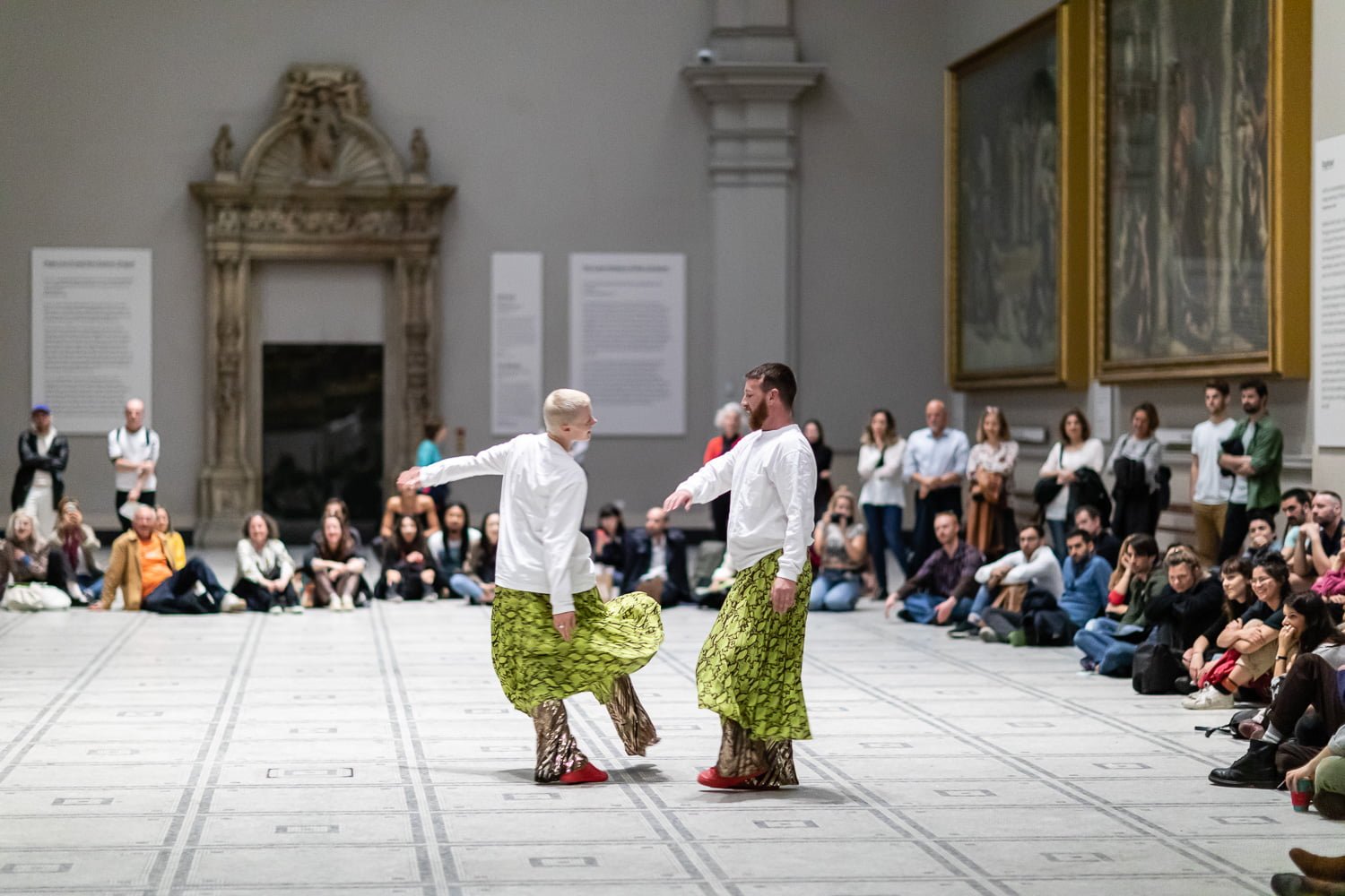 Two dancers wearing white long sleeved tops and a bright yellow skirt with a black pattern dance in the middle of an art gallery, surrounded by an audience of people on all sides.