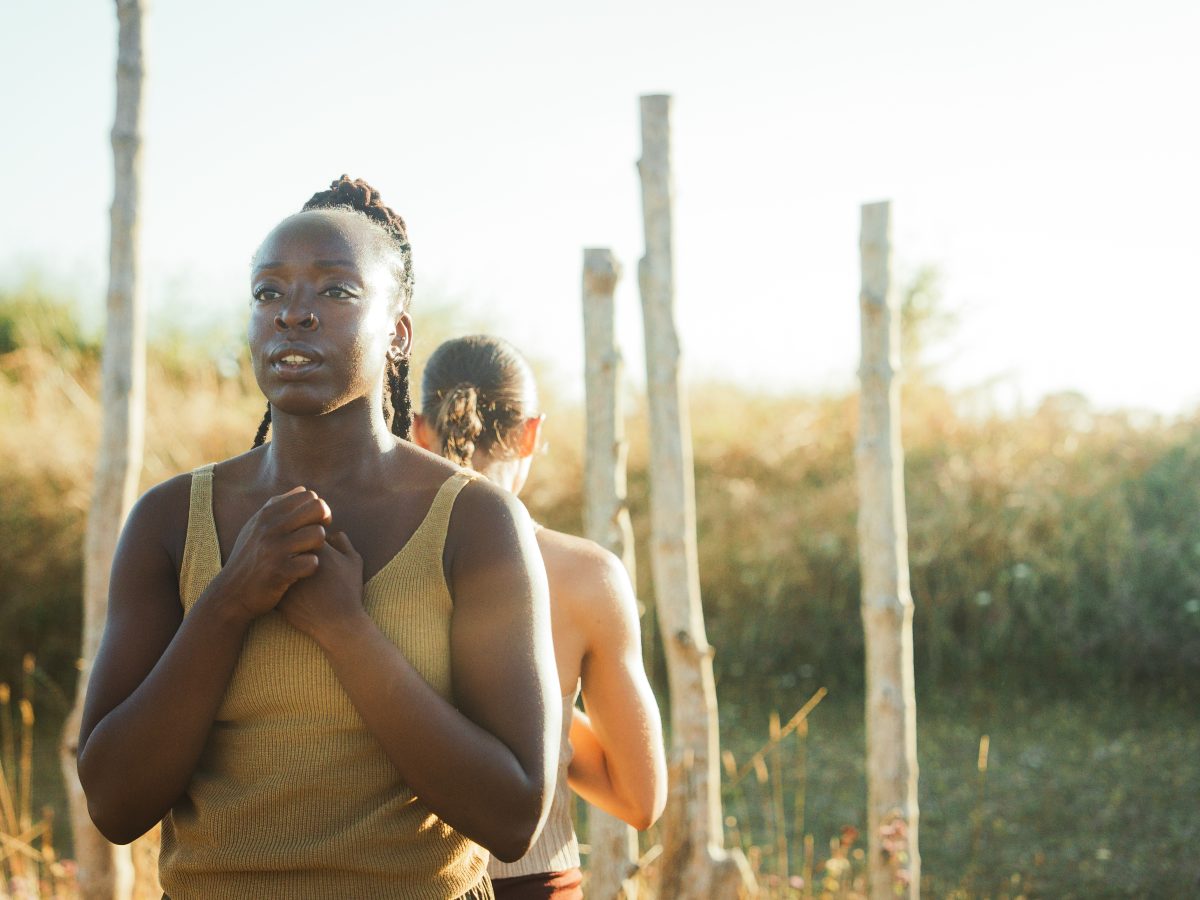 Two dancers stand back to back, their arms clasped gently in front of their hearts. They stand in front of a series of wooden posts standing tall at the centre of a Bronze Age pond barrow.