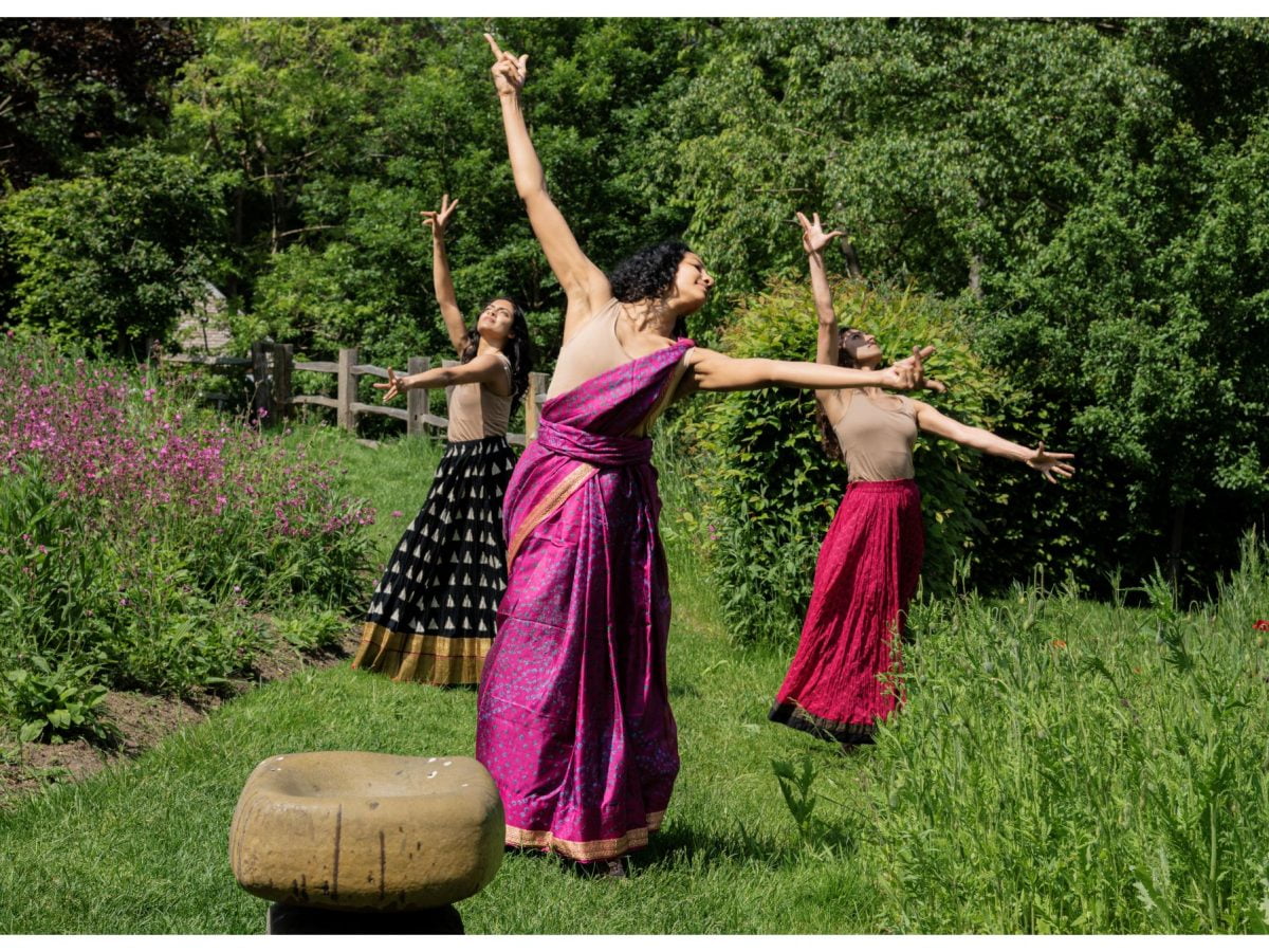 3 dancers move with arms outstretched in a garden, with lush green flowers and trees all around.