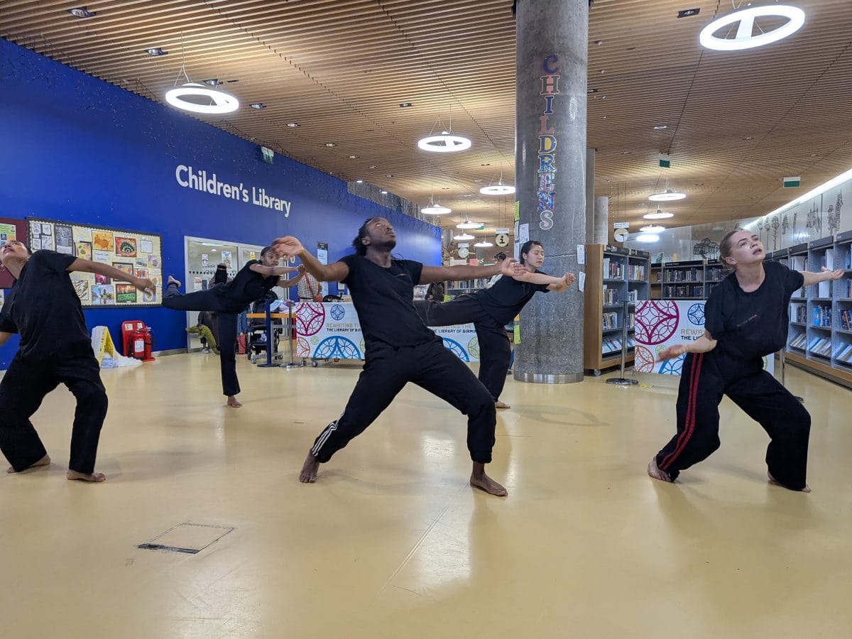 Five dancers dressed in black perform in a library space